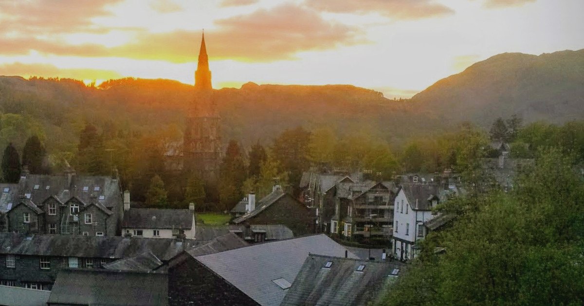 Sunset over Ambleside. In the foreground, the slate-grey houses of the village. In the middle-ground, a church, glowing from the setting sun behind, just going down below the mountains in the background.