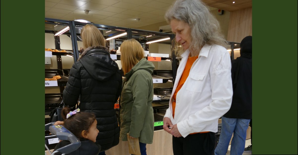 Lena in a white jacket and black jeans, standing in a shop looking down at a little girl, who is looking up at her in response. A couple of shoppers are at the bakery section behind them, with their backs turned as they select their shopping. Probably one of them is the little girl's mother.