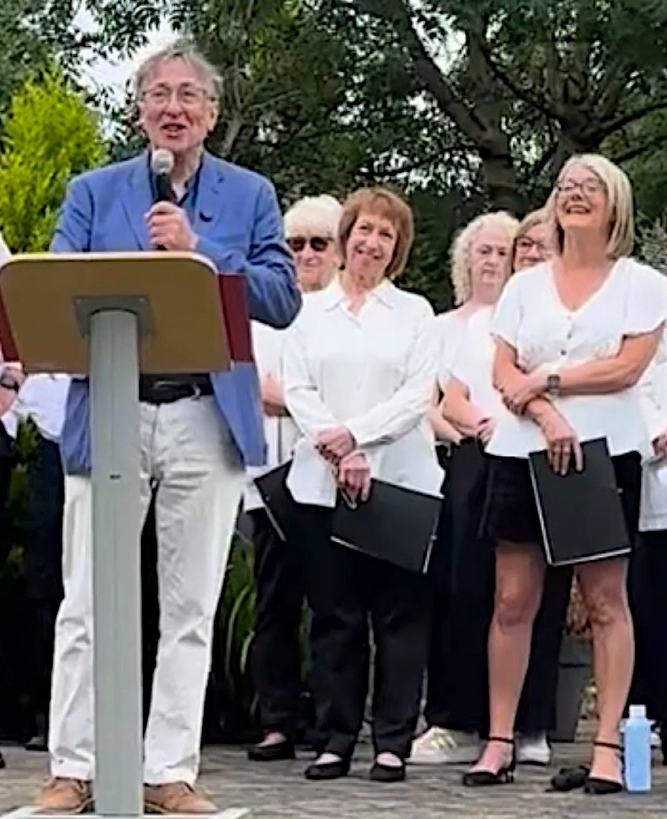 Alan Miles speaking to an audience at the Moya Cole Hospice in May 2025. Behind him a choir is standing, laughing.